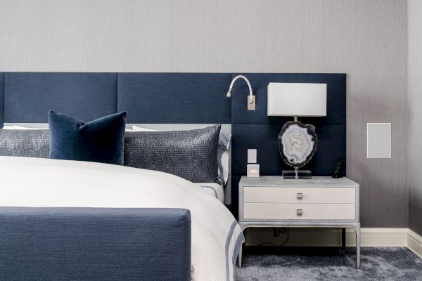 A modern, minimalistic bedroom with a navy blue and white color scheme, showcasing a bed, pillows, nightstand, lamp, and a clock.