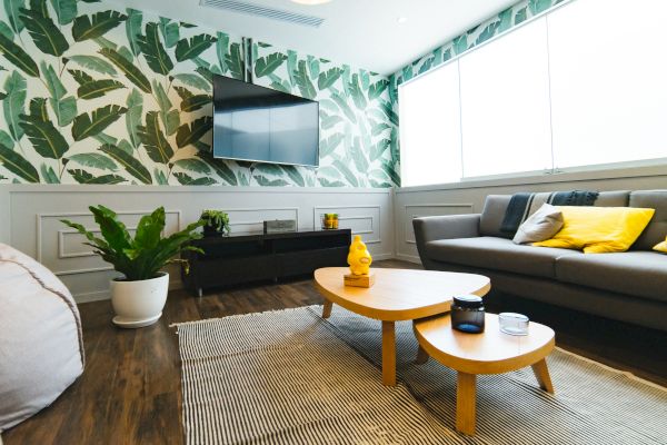 A modern living room features a tropical leaf wall, a mounted TV, a grey sofa with yellow pillows, wooden tables, plants, and large windows.