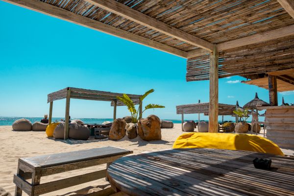 A beachfront setting with shaded wooden structures, seating areas, and sandy surface under a bright blue sky, offering a relaxing vibe.