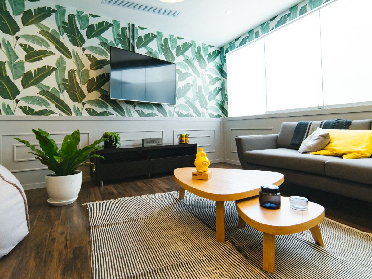 A modern living room with a tropical leaf-patterned accent wall, a grey couch with yellow cushions, a TV, and a wooden coffee table on a striped rug.