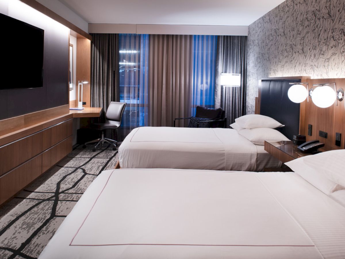 A modern hotel room features two neatly made beds, a flat-screen TV, a desk with a chair, and contemporary decor with a patterned accent wall.