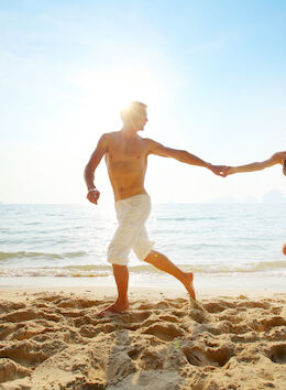 A couple is running hand in hand on a sandy beach with the ocean in the background and the sun shining brightly.