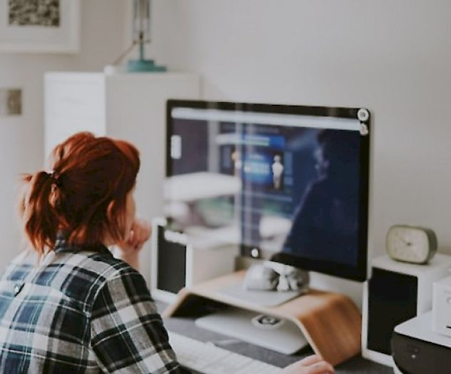 A person with red hair sits at a desk, working on a computer. There are various office supplies and devices around them.