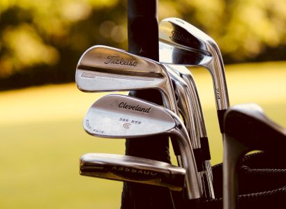 The image shows a set of golf irons from brands Titleist and Cleveland, placed in a golf bag with a blurred background.