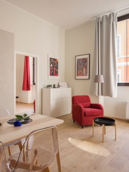 The image shows a bright living room and kitchen area with a dining table, red armchair, sofa, and large window overlooking a colorful building.