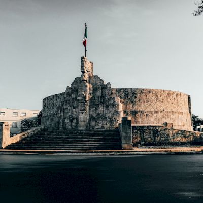 A circular stone monument with stairs and a flag on top, set against a quiet street and clear sky backdrop.