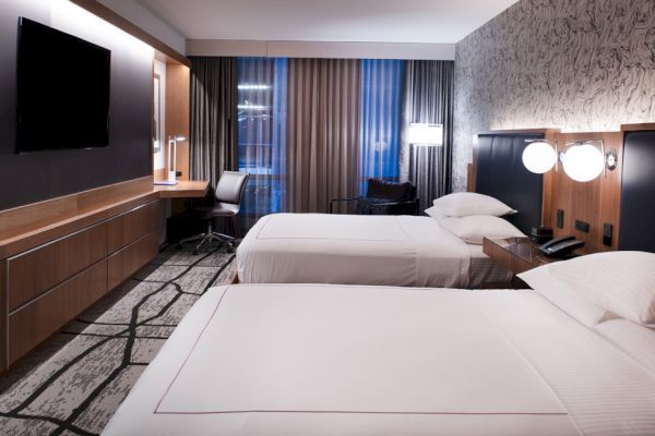 A modern hotel room with two beds, a TV, a desk with a chair, and stylish decor, including wall art and lighting, in a sleek, cozy setting.