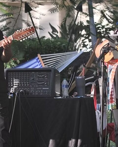 Two musicians perform outdoors with guitars and microphones, surrounded by audio equipment and greenery.