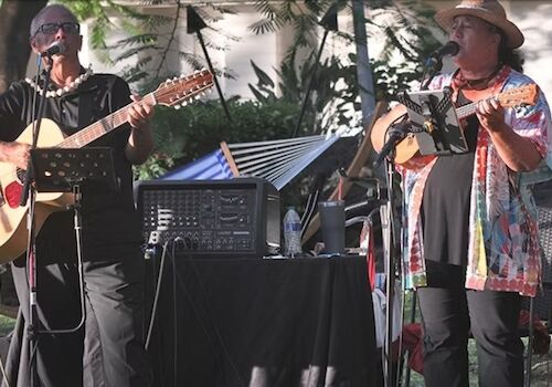Two musicians perform outdoors with guitars and microphones, surrounded by audio equipment and greenery.