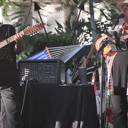 Two musicians perform outdoors with guitars and microphones, surrounded by audio equipment and greenery.