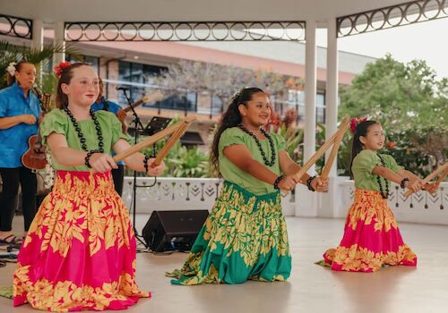Three dancers in colorful skirts perform with wooden sticks on stage, accompanied by a musician.