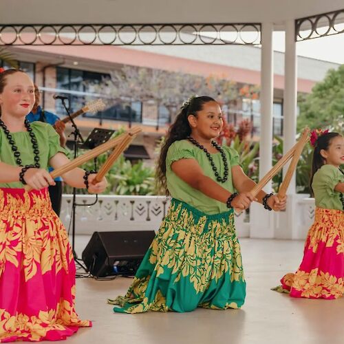 Three dancers in colorful skirts perform with wooden sticks on stage, accompanied by a musician.