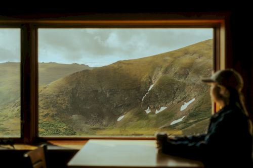 A person seen in silhouette with a hat, sitting inside, looking out at a mountainous landscape through a large window.