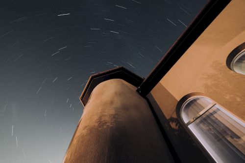 The image shows a night sky with star trails captured above a tall building structure, with windows and architectural details visible.