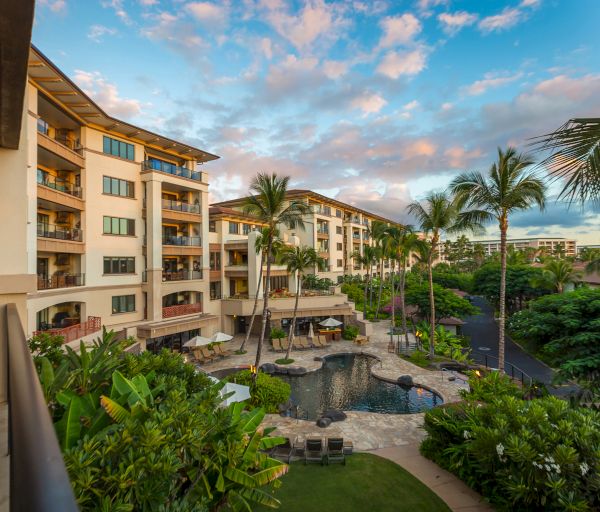 The image shows a resort with multiple buildings, a pool area, palm trees, lush greenery, and a vibrant sky at sunset.