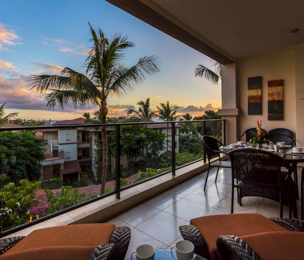 A balcony with a dining table, overlooking a tropical landscape with palm trees and residential buildings, captured during sunset.