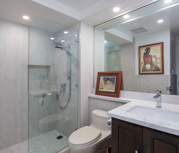 This image shows a modern bathroom with a glass-enclosed shower, a toilet, a sink on a marble countertop, a large mirror, and framed artwork.