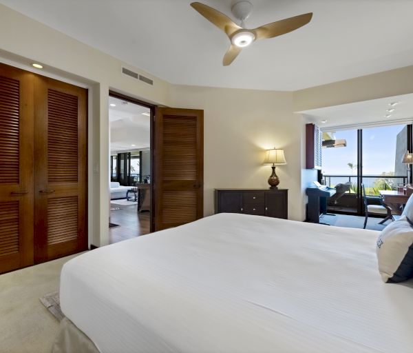 A modern bedroom with a double bed, ceiling fan, wooden closet doors, a nightstand with a lamp, and a seating area near large windows with an ocean view.