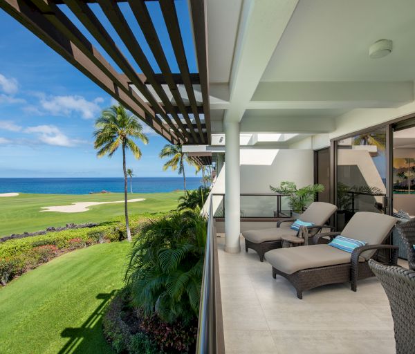 A patio with lounge chairs overlooks a green golf course and the ocean, with palm trees under a partly cloudy blue sky.