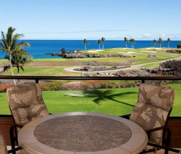 The image shows a patio with a round table and four cushioned chairs, overlooking a scenic golf course, palm trees, and a view of the ocean.