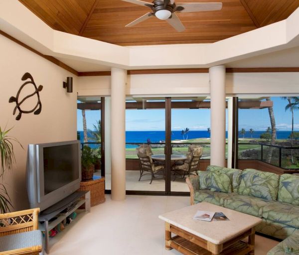 A living room with ocean views, tropical-themed decor, plants, a TV, wicker furniture, and a patterned sofa. A wall art of a turtle is displayed.