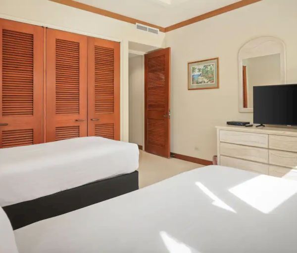 A neatly arranged bedroom with two beds, a TV on a dresser, wooden closets, a lamp, and framed art on the walls, with natural light coming in.