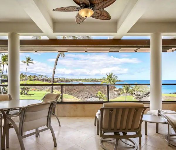 A covered patio with seating and a ceiling fan overlooks a scenic coastal view with palm trees, the ocean, and a clear blue sky in the background.