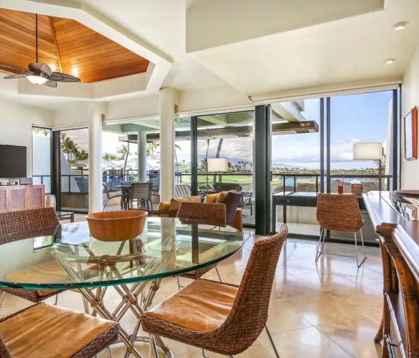 This image shows a modern, open-plan living and dining area with a glass table, wicker chairs, and large windows providing a scenic view.