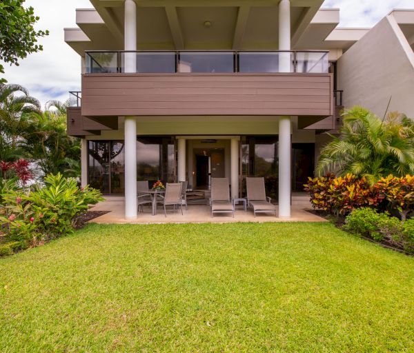 The image shows an outdoor patio area of a modern house with a green lawn, outdoor seating, and tropical plants surrounding the space.