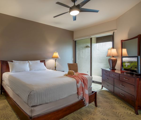 A cozy bedroom features a double bed, wooden furniture, two lamps, a TV, a ceiling fan, and sliding glass doors with a view outside.