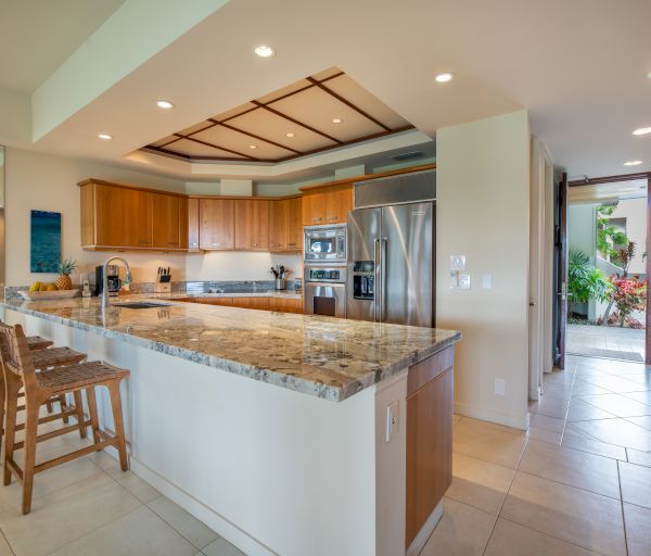 A modern kitchen with a marble island, bar stools, stainless steel appliances, wooden cabinets, and tile flooring opens to an outdoor area.