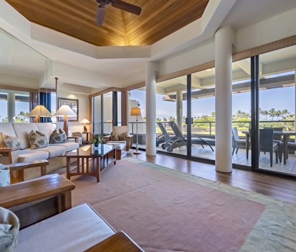 A spacious and elegantly decorated living room with large windows opening to a balcony with outdoor seating and a scenic view.