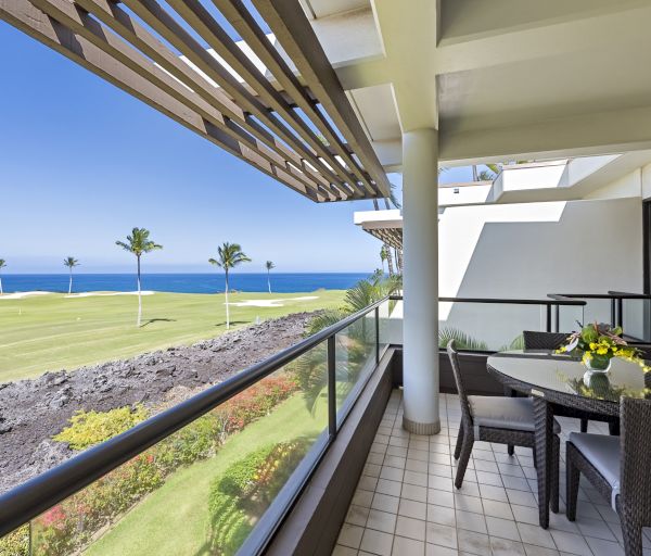 A balcony with a table and chairs, overlooking a golf course and ocean with palm trees in the distance, on a sunny day.