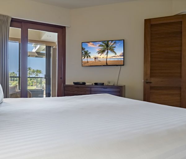 A cozy bedroom features a large bed, a wall-mounted TV displaying a beach scene, and a balcony with outdoor views through glass doors.