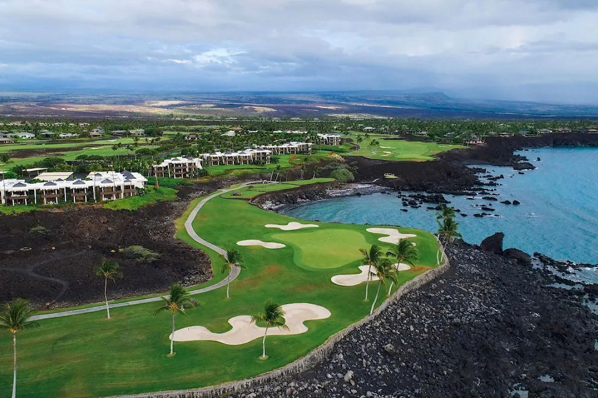 Aerial view of a coastal golf course with green fairways, sand bunkers, palm trees, and adjacent modern buildings, near a rocky shoreline and ocean.
