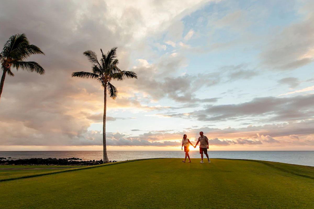 A couple holding hands walks on a grassy area with palm trees, overlooking a serene ocean and a vibrant sunset sky.