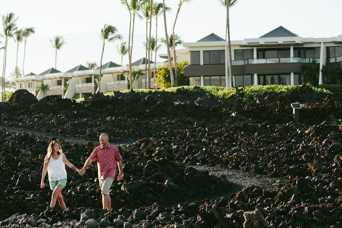 A couple holds hands while walking on rocky terrain near palm trees and resort buildings in the background.