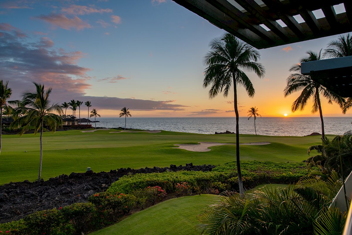 A scenic view of a sunset over a golf course near the ocean, with palm trees and lush greenery in the foreground, and a calm sky.