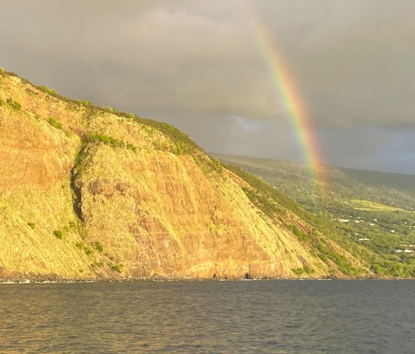 The image shows a coastal cliffside with a rainbow arcing over it. The sky is partly cloudy, and the water in the foreground is calm.