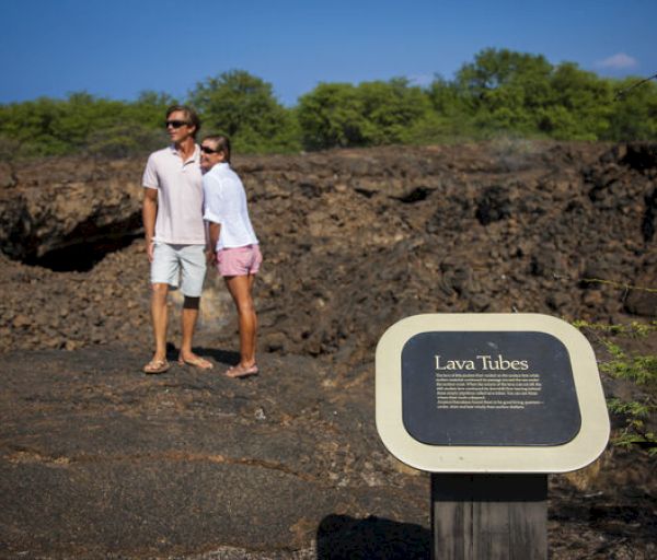 A couple stands near a "Lava Tubes" sign in a rocky landscape with green foliage under a clear blue sky.