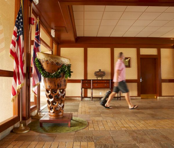 A person in casual attire walks through a lobby with flags, a large decorative object, and wooden furniture.