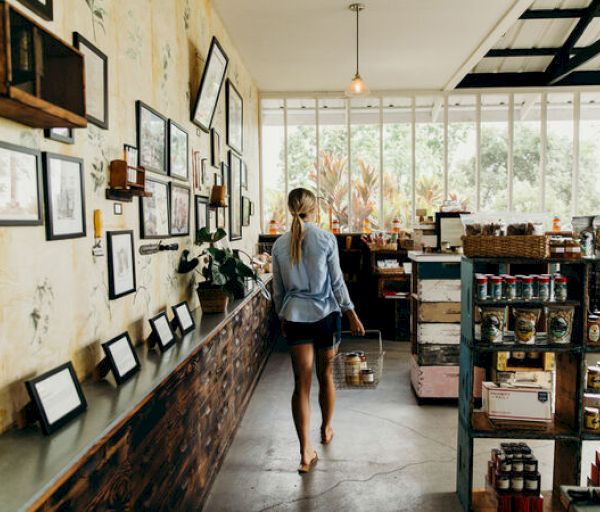 A person walks barefoot in a cozy store with framed pictures on the wall, shelves stocked with items, and warm lighting. The sentence ends here.