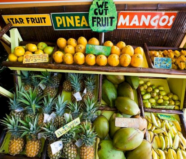 The image shows a fruit stand with star fruits, pineapples, mangoes, oranges, bananas, coconuts, and limes, with a sign saying "WE CUT FRUIT."
