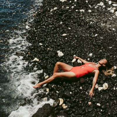 A person in an orange swimsuit is lying on a rocky beach with waves gently reaching their feet, surrounded by black pebbles and a few larger rocks.