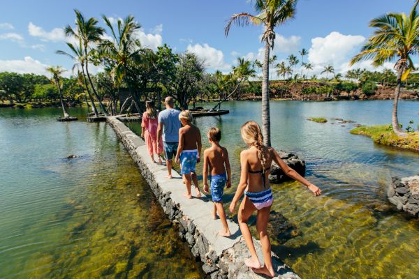 A group of people, including adults and children, walk along a narrow stone path over a body of water with palm trees and green vegetation around.