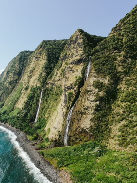 This image features a scenic coastal cliff with multiple waterfalls cascading down into the ocean, surrounded by lush greenery under a clear sky.