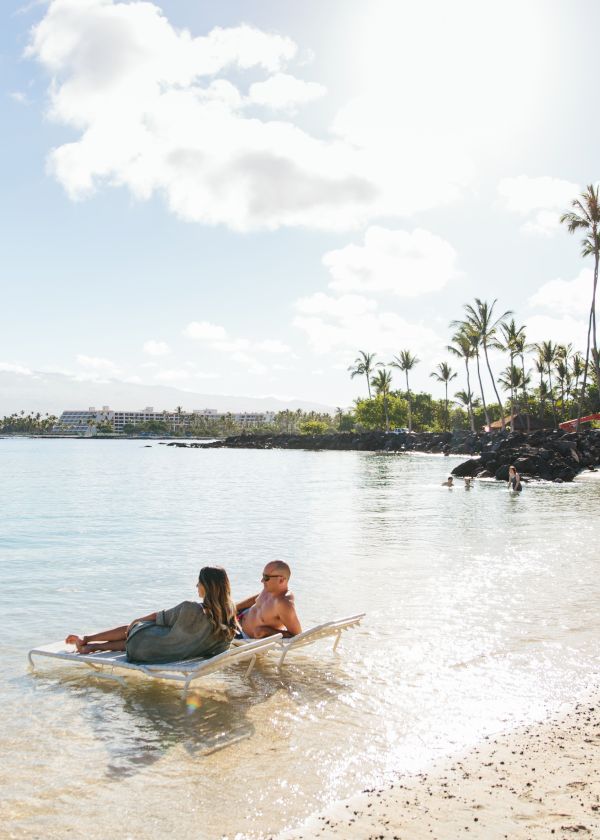 Two people relax on a lounge chair partially submerged in the shallow, clear water of a tropical beach with palm trees and a calm shore.