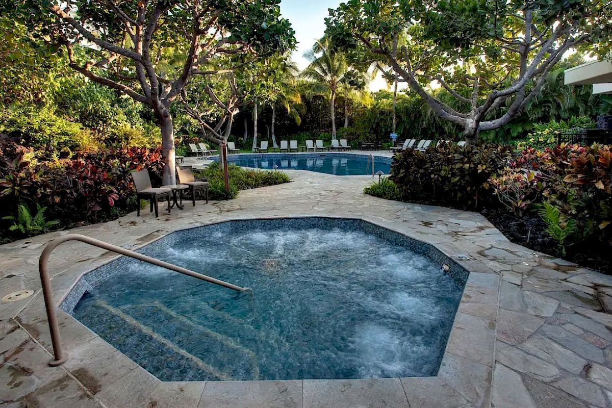 A serene outdoor pool area with a hot tub in the foreground, surrounded by lush greenery and trees, perfect for relaxation.