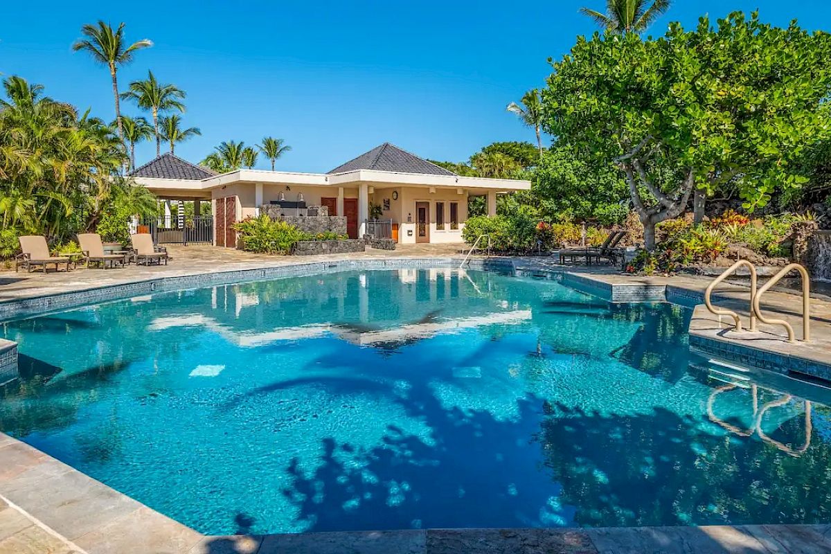 A serene outdoor pool surrounded by lounge chairs, palm trees, and a building under a clear blue sky.
