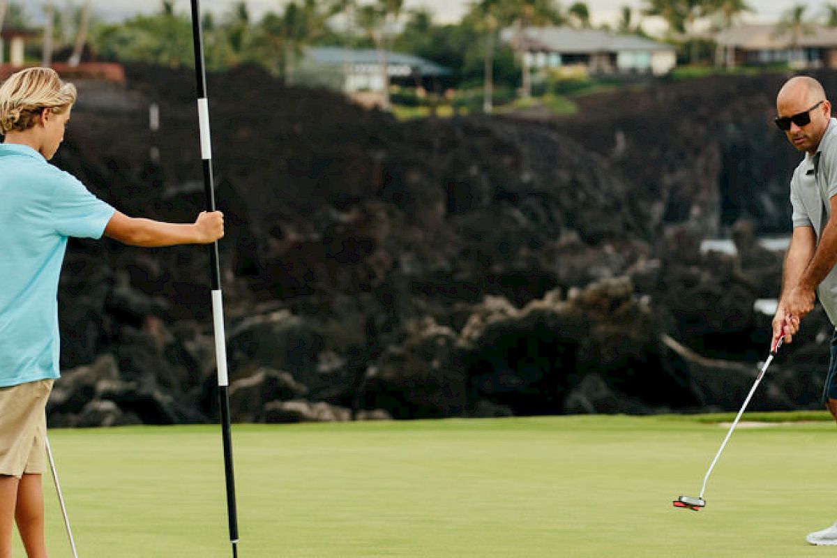 Two people are playing golf; one is putting while the other holds the flagstick near a rocky coastal background.